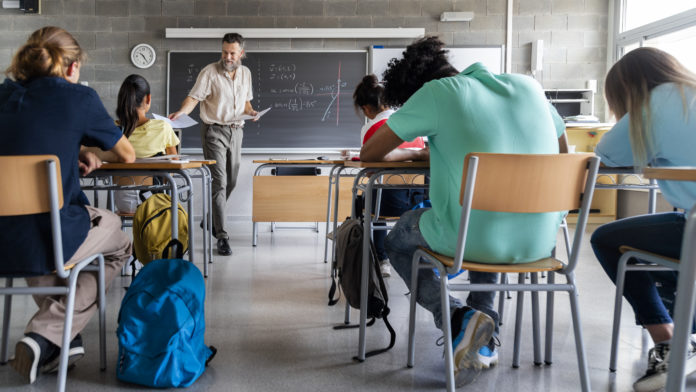 Mature caucasian man teacher hands out exams to multiracial high school students. Students ready to take exam. Horizontal photo banner