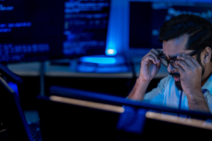 South Asian male developer concentrates silhouetted against bright monitor screens showing programming interfaces in a dark tech lab.